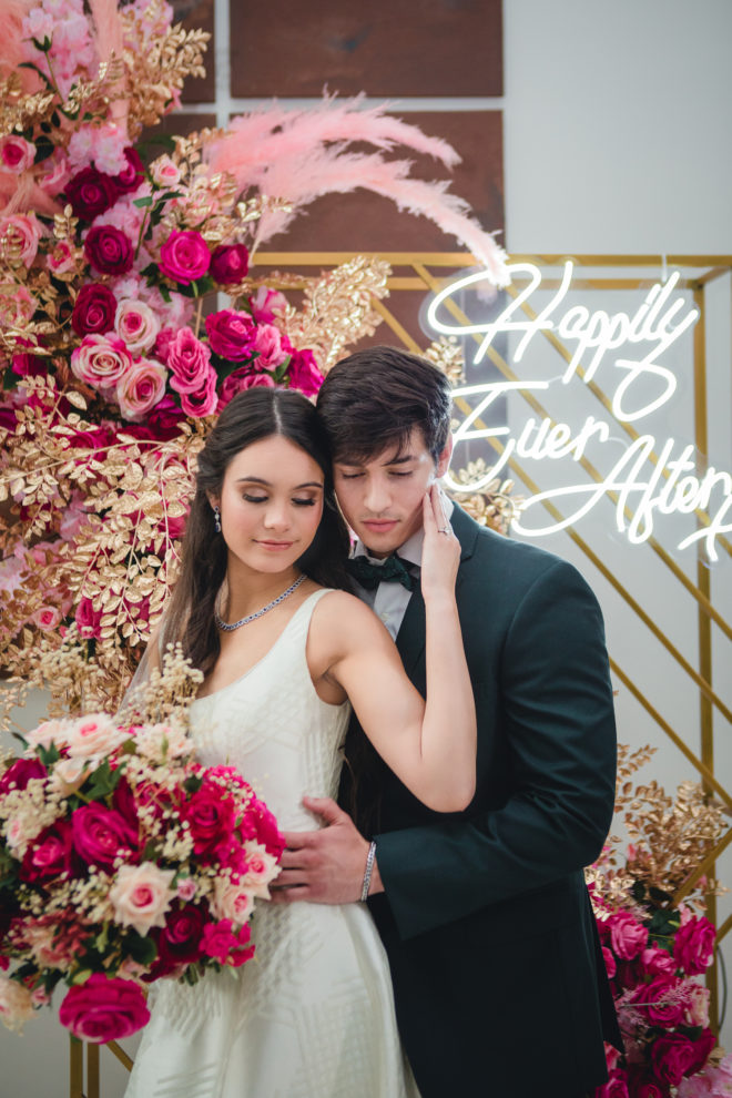 Bride holding groom's face with gold background, lush hot pink and blush florals and lit LED sign which reads, "Happily Ever After".