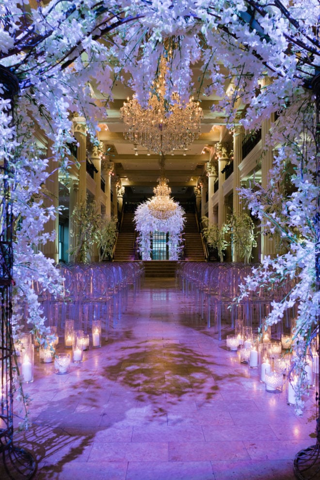 Wisteria covered arch leading to ceremony aisle with glowing candles, lavender colored uplighting and white acrylic ghost chairs designed by Plants N' Petals at the Corinithian Houston.