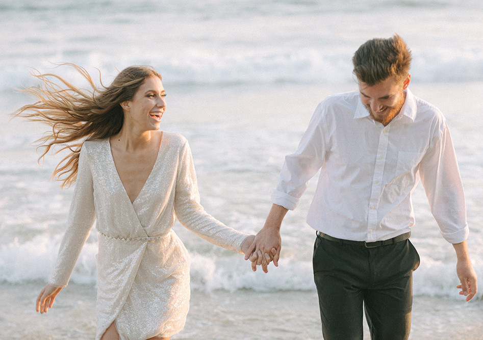 A candid photo of a bride and groom as they hold hands and walk in the sand with the ocean in the background.