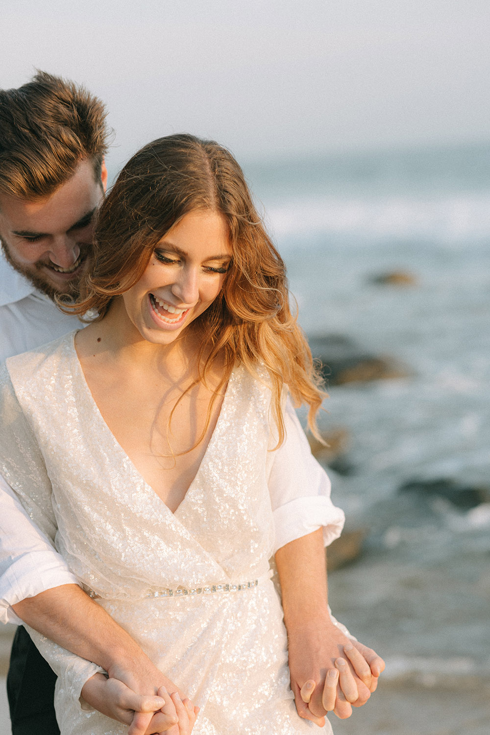 Man embraces a woman in a sequin mini dress on sunny Orange County beach with the waves of the Pacific Ocean behind them. 