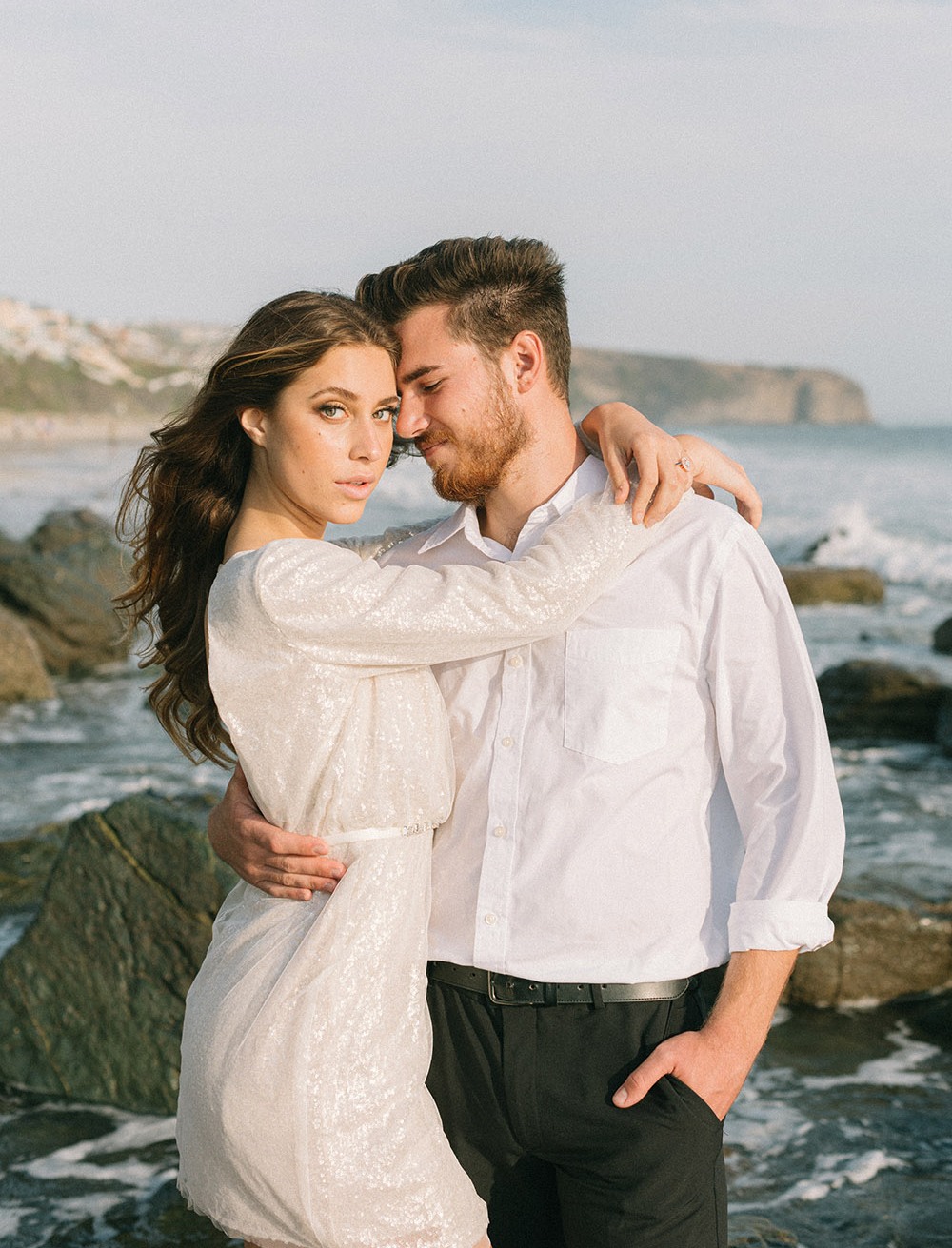 The couple in this beach elopement styled shoot embrace each other on the beach as the sunlight illuminates the bride's draped sequined mini dress.