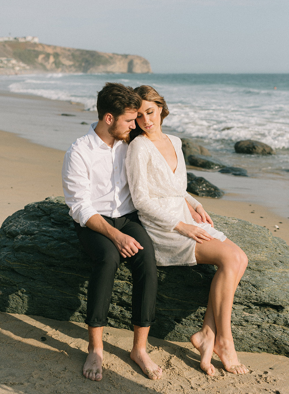 A groom in a white button down and black pants sits next to his bride in a stylish sequined mini dress by Vera Wang during this beach elopement shoot.