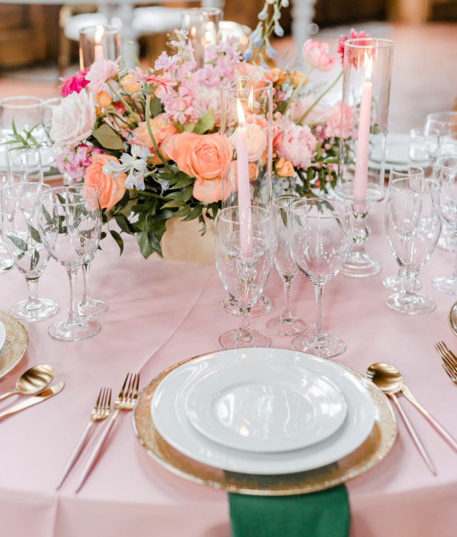 Round table with pink tablecloth set with white and gold placesettings, dark green napkins, lit pink tapered candles and a fresh floral centerpiece with garden roses, peonies and greenery inside a reception space at The Carriage House. 
