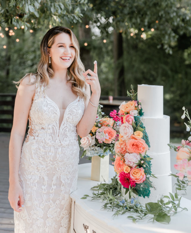 Woman in white fitted lace with plunging illusion neckline smiling in the distance while holding up a finger covered in icing next to a four tiered white wedding cake adorned with fresh florals atop a vintage dresser on the patio of the Carriage House wedding venue at a wedding styled shoot by Amy Maddox Photography. 