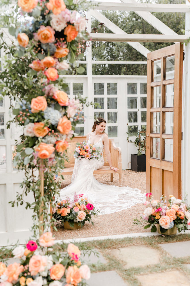 Woman in white fitted gown sitting on antique pew inside white structure adorned with fresh orange and pink florals and greenery at a wedding styled shoot by Amy Maddox Photography at The Carriage House in Conroe, Texas.