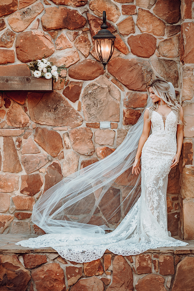 A bride poses in front of a terra cotta stonework wall wearing a lace dress and a cathedral length veil.