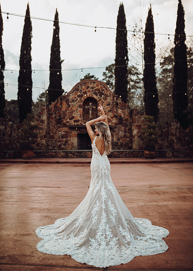 A bride stands in front of the Grand Fireplace at Madera Estates wearing her lace wedding gown with plunging back and long, scalloped train.
