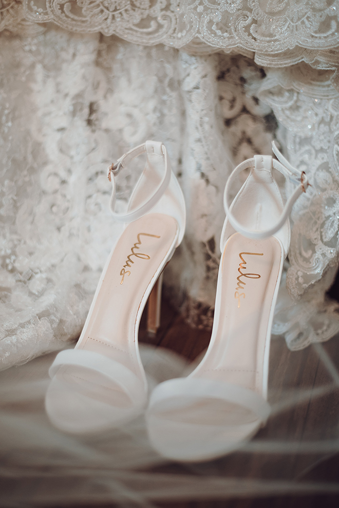 Close up of white Lulus heeled strappy sandals resting on the ground surrounded by the lace train of the bride's wedding dress.