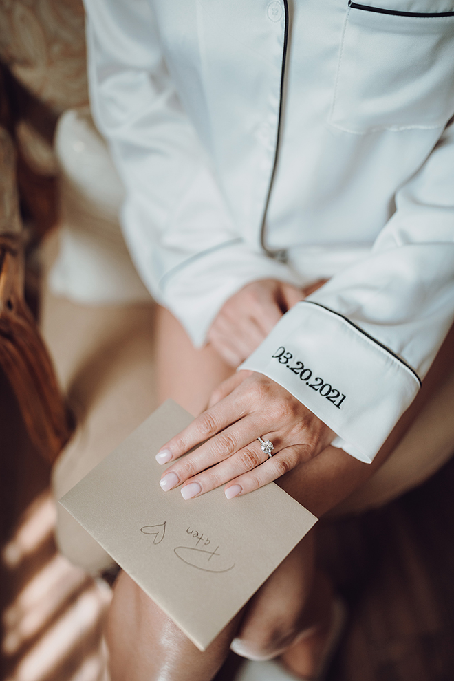 A bride in white pajamas with 03.20.2021 embroidered on her sleeve holds a letter in a brown envelope for her soon-to-be husband. 