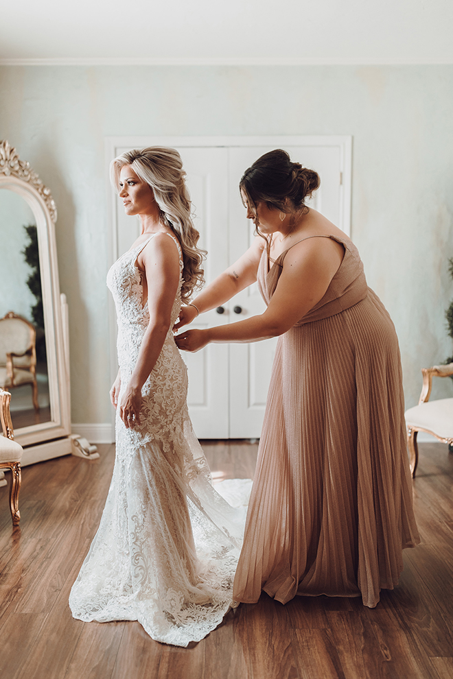 A bridesmaid in a floor-length rose gold dress helps button the brides lace wedding dress. 