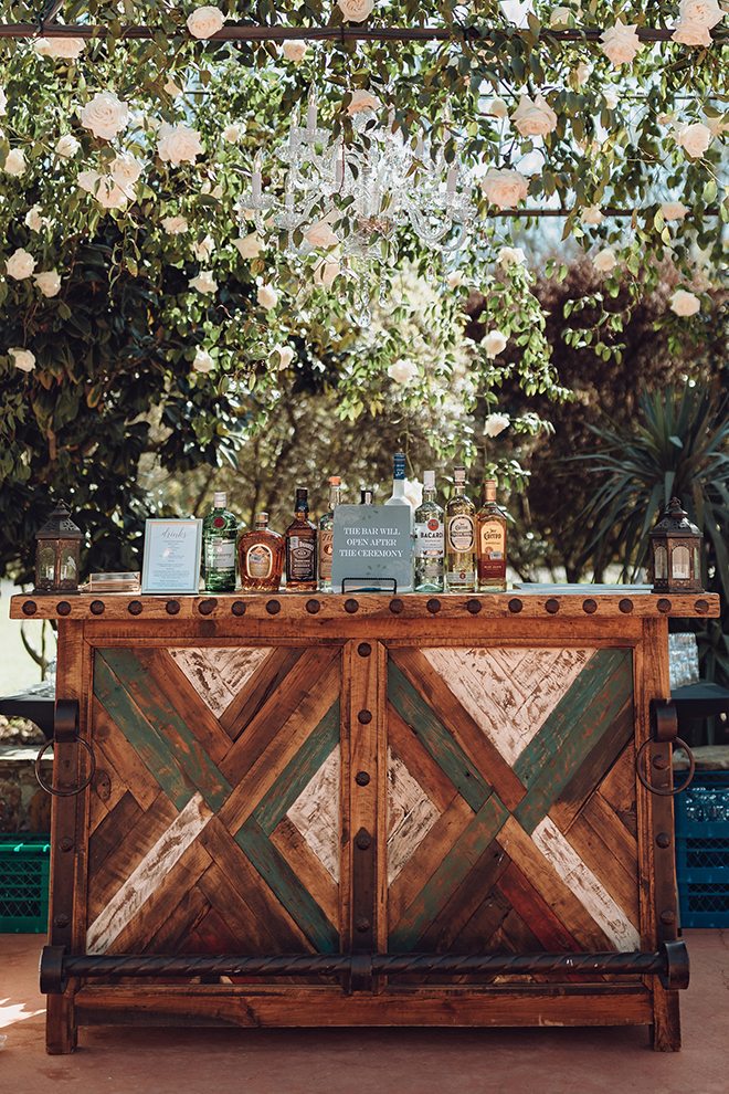 A rustic bar cart sits under a canopy of white and blush roses and cascading greenery, unopened until after the wedding ceremony.