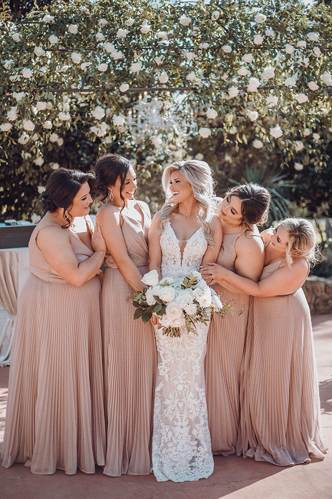 A blonde, smiling bride in a lace wedding dress holds her bouquet surrounded by her bridesmaids in floor-length blush dresses.