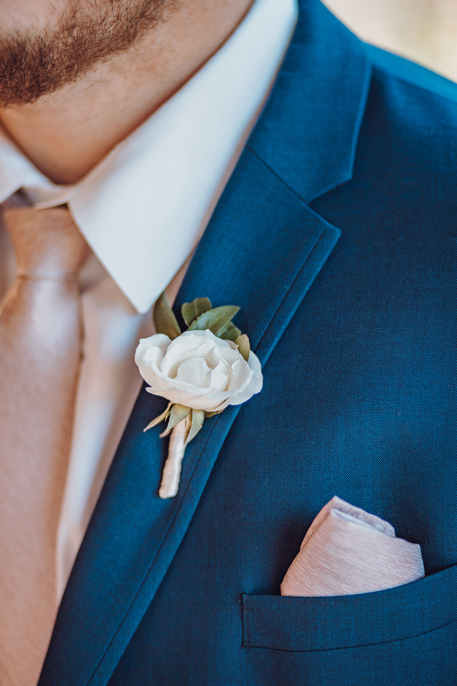 A close up shot of a grooms white boutonniere against a navy blue suit with rose gold accents.