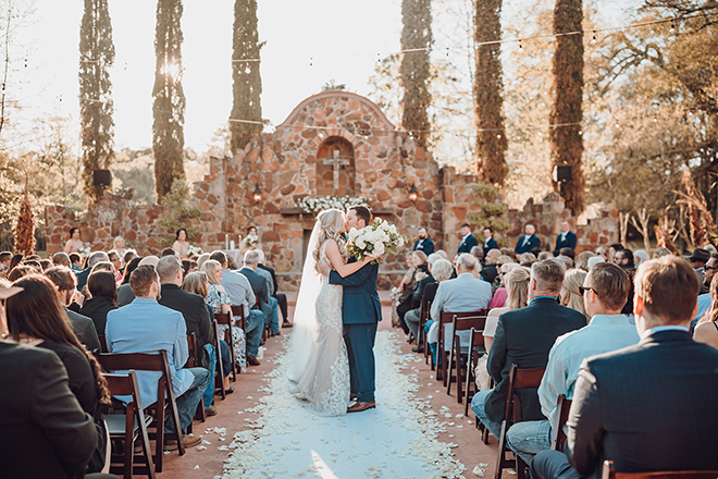  A newlywed couple kiss in the aisle after their wedding ceremony at the southwestern-inspired Madera Estates Houston wedding venue.