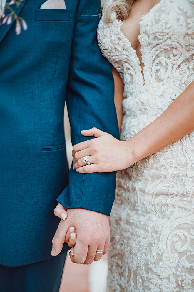 A bride in a plunging lace wedding dress holds the hand of her groom in a navy blue suit. 