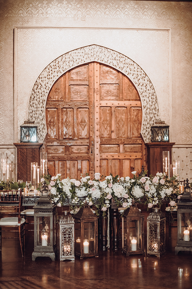 Candle-lit lanterns illuminate a full row of white and blush wedding florals at the bride and grooms table at the white and rose gold wedding reception.