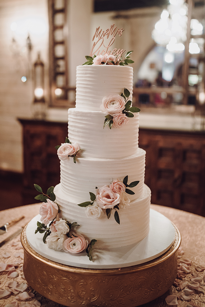 A traditional, white, 4-tiered wedding cake is adorned with blush roses and a rose gold, cursive Mr. and Mrs. Richey cake topper.