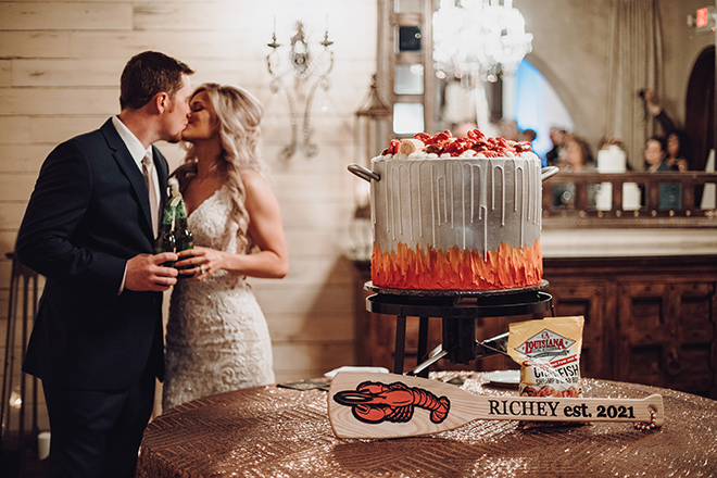 The bride and groom kiss in front of a crawfish broil themed grooms cake at their white and rose gold wedding reception.