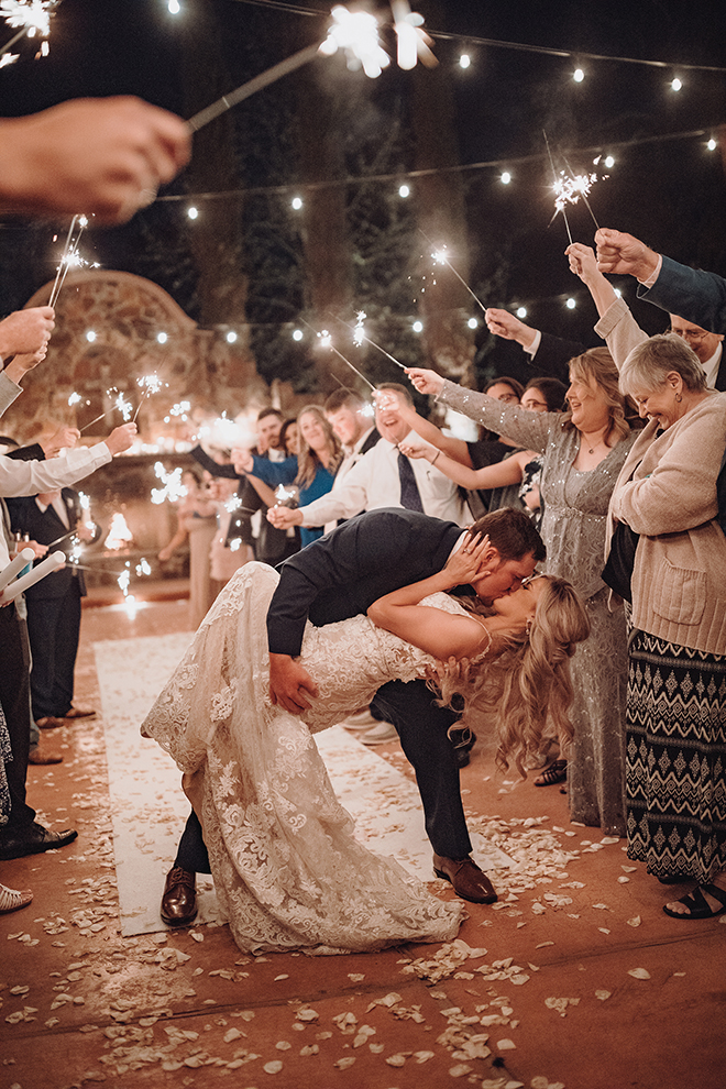 A groom lowers his bride and gives her a kiss under a canopy of sparklers held by their wedding guests.