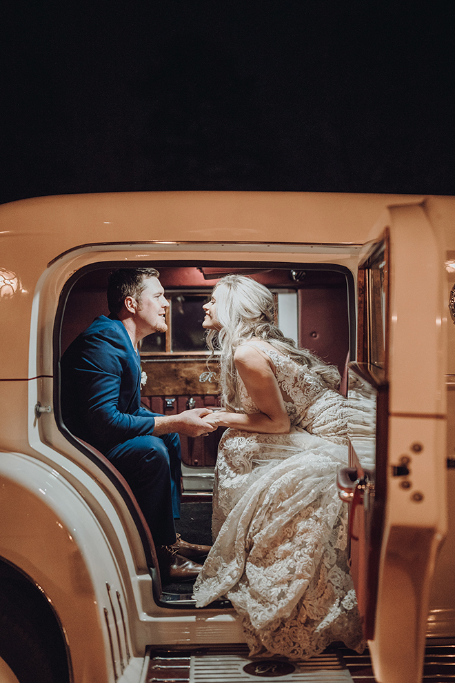A bride and groom gaze into eachothers eyes while posing inside a vintage white car. 