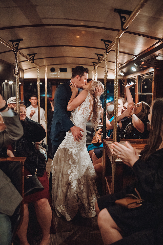 A newlywed couple kisses inside a trolley car after their wedding while their wedding guests look on. 