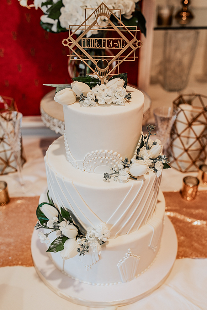 A wedding cake with 1920's-esque glamour is adorned with geometric white sculpted icing and topped with a vintage-meets-modern cake topper and traditional white florals. 
