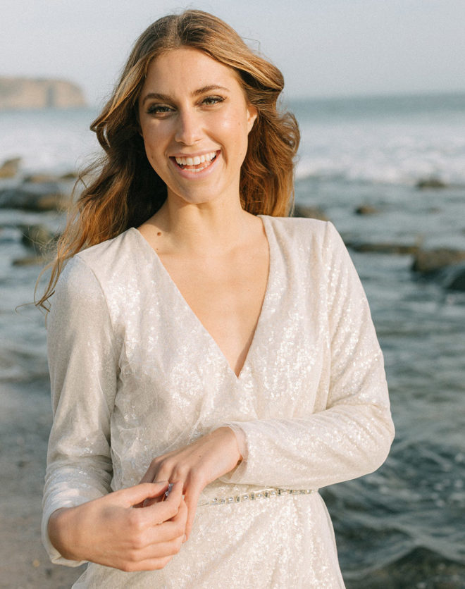 Smiling woman with wavy hair and sequin white long sleeve mini dress with sparkling belt on a beach in California during an elopement editorial by Esther Makau Photography. 