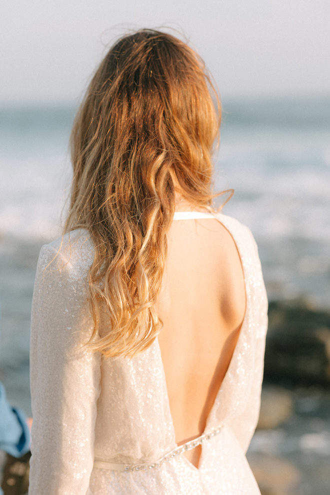 Woman facing Pacific ocean wearing a deep v-back mini sequin dress by Vera Wang on an Orange County California beach during an elopement shoot by Esther Makau Photography. 
