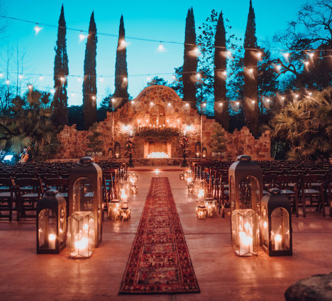 Ceremony set-up in Madera Estate's outdoor garden. A red persian rug serves as the aisle, while an ecclectic mix of bronze lanterns with glowing candles flank to the sides, leading up to a rustic stone fireplace adorned with glowing candles, blooming florals and additional lanterns. Glowing string lights hang above the ceremony chairs in an al fresco ceremony scene at wedding venue, Madera Estates. 