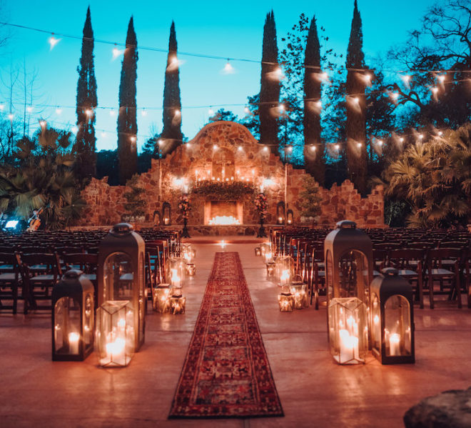 Ceremony set-up in Madera Estate's outdoor garden. A red persian rug serves as the aisle, while an ecclectic mix of bronze lanterns with glowing candles flank to the sides, leading up to a rustic stone fireplace adorned with glowing candles, blooming florals and additional lanterns. Glowing string lights hang above the ceremony chairs in an al fresco ceremony scene at wedding venue, Madera Estates.