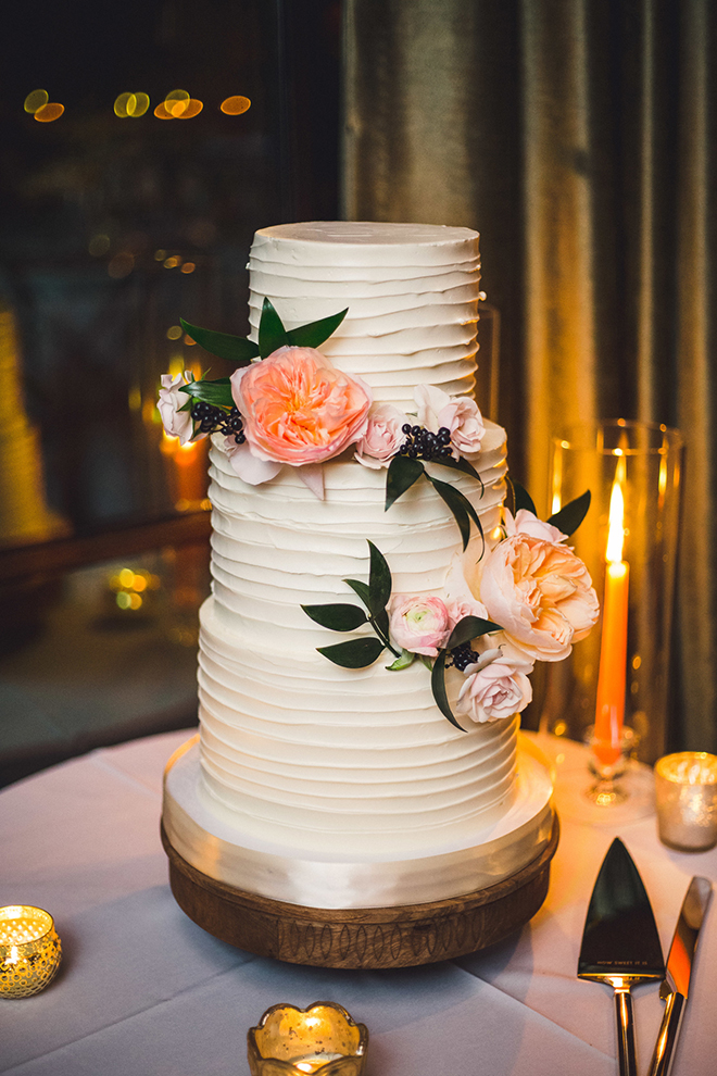 A three-tier spatula iced cake with ivory frosting sits atop a rustic wood cake platform and is adorned with peach florals and blue berries.