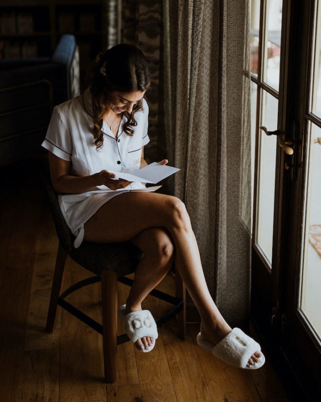 Bride, in white pajamas with monogrammed pocket and white fuzzy slippers that say "I do", reads a card by the light of a hotel suite window. 