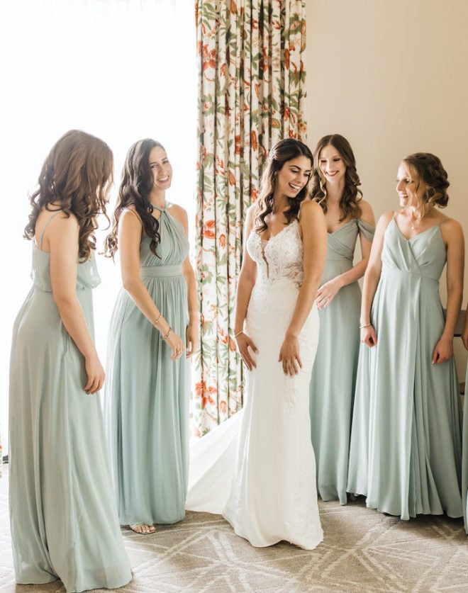 Bride, in a white fitted wedding gown, showing four bridesmaids, wearing sage green dresses, the back of her wedding gown in a hotel room suite at the Hyatt Regency Lost Pines Resort and Spa. 