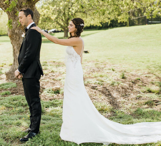Groom, in black suit, with his back to the bride, in strappy white gown with long train, who has her hand on his shoulder during the first look on the lawn of Hyatt Lost Pines Resort. 