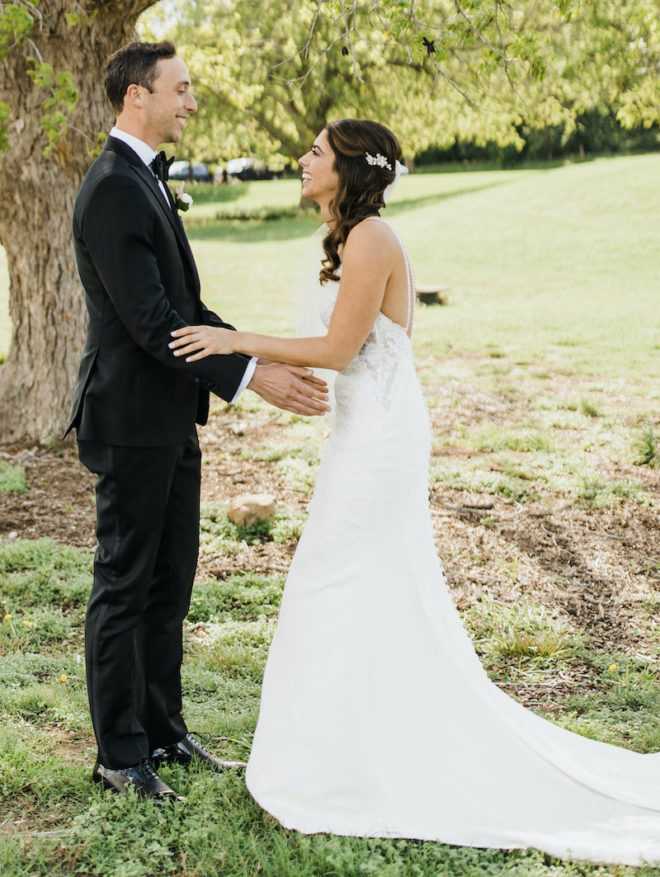 Groom holds out arms for an embrace while bride, wearing long white gown, smiles at him on the lawn of Hyatt Lost Pines Resort located in Bastrop, Texas. 