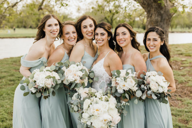 Bride, holding blooming white bouquet, smiling with five bridesmaids in sage green long dresses next to the Colorado River at Hyatt Regency Lost Pines Resort and Spa. 