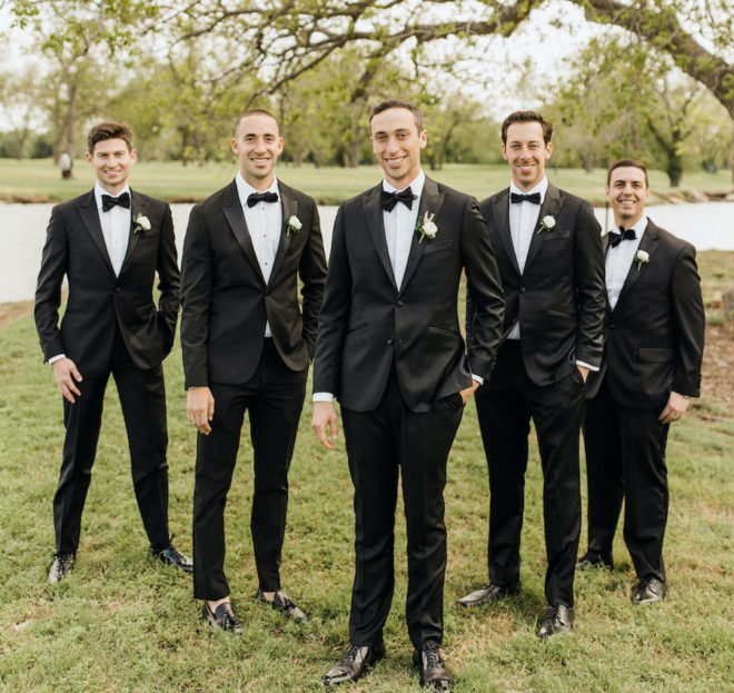 Four groomsmen in black tuxedos and black bowties standing behind a smiling groom on the green lawn next to a river at Hyatt Lost Pines Resort near Bastrop, Texas. 
