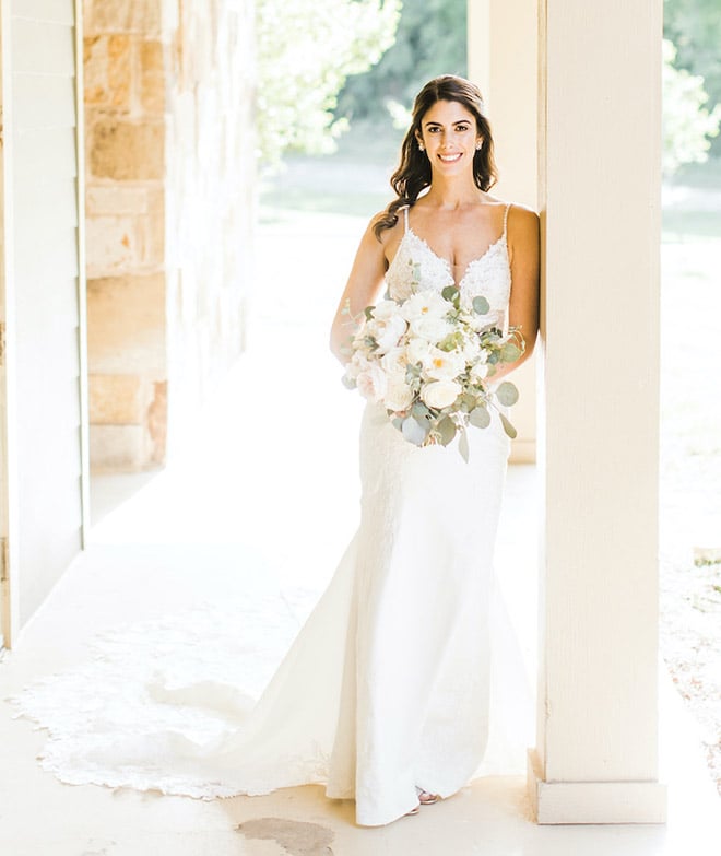 Bride, in beaded white wedding gown with long lace train, holds lush white florals with sage colored greenery, on a covered patio at Hyatt Regency Lost Pines Resort and Spa. 