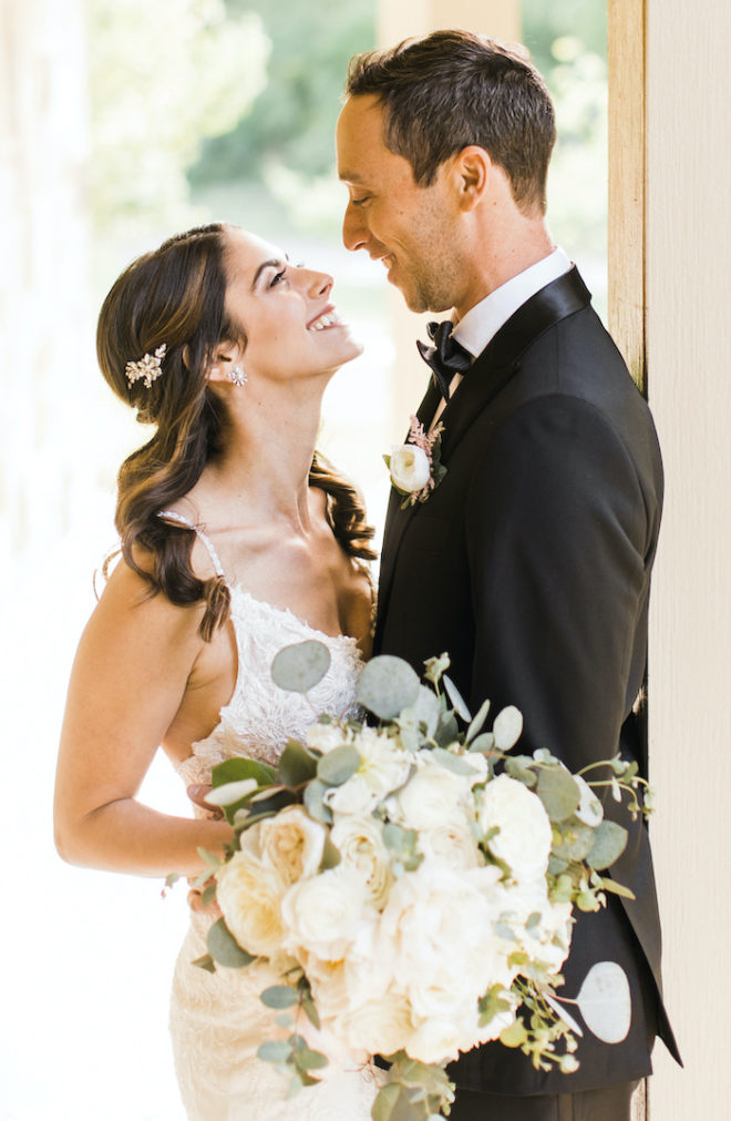 Bride, in beaded white wedding gown holding blooming florals, holds blooming white floral bouquet while holding the smiling groom on the outdoor porch at Hyatt Regency Lost Pines Resort and Spa. 