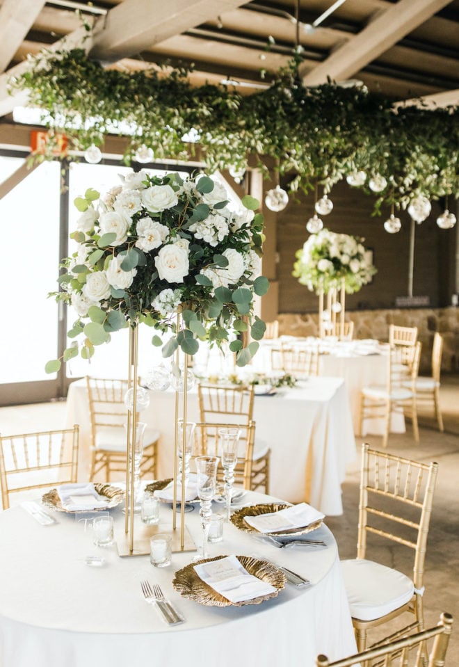 Banquet tables adorned with gold chivari chairs, tall blooming white floral centerpieces and gold plate charges amidst lush hanging greenery with hanging glass globes in the interior event space of Hyatt Lost Pines Resort. 