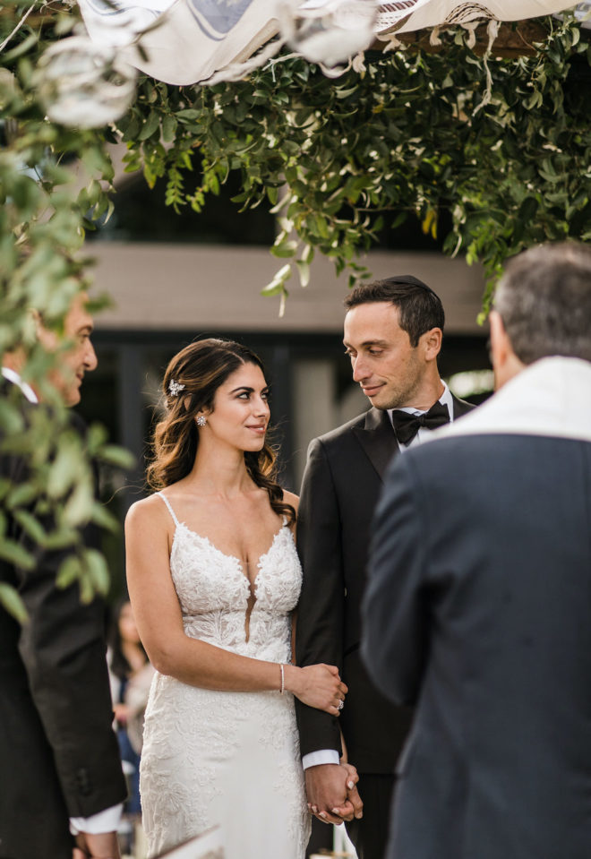 Bride, in beaded fitted white gown and half updo, smiles at the groom, in an all black tuxedo and black bowtie, during an alfresco ceremony underneath a greenery and floral chuppah on the LBJ pavilion patio at Hyatt Regency Lost Pines Resort and Spa. 