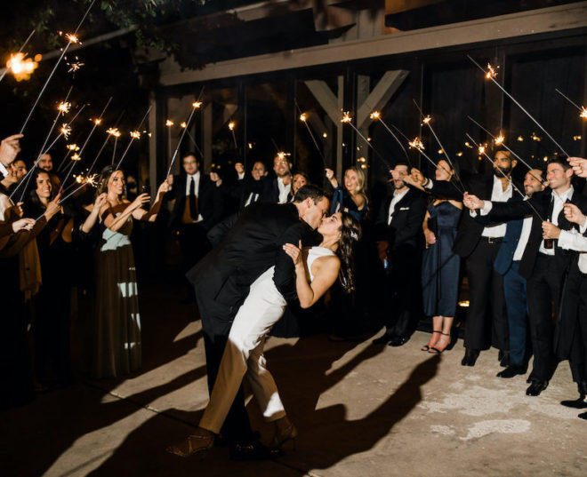 Groom, in all black tuxedo, kisses bride, in white bridal jumpsuit, during a sparkler send-off on the LBJ pavilion patio at Hyatt Regency Lost Pines Resort and Spa. 