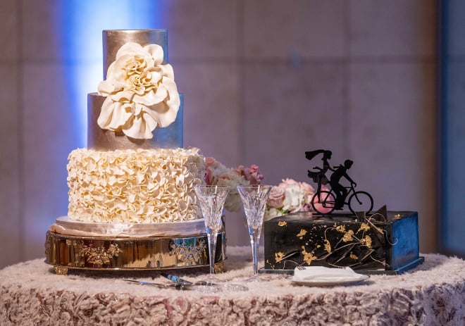 A white and gold wedding cake sits next to a striking single-tier square groom's cake in black with gold foil accents, topped with the silhouette of a couple on a bicycle by Common Bond Bistro & Bakery..