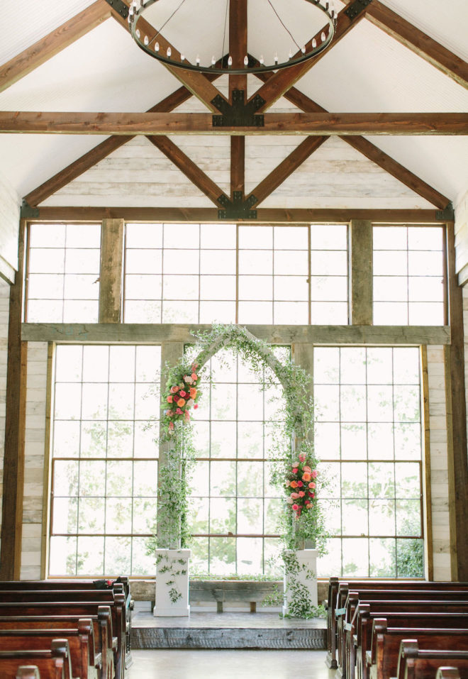 Sun dappled ceremony space with large industrial windows, aisles of wood pews, industrial chandelier overhead and a floral and greenery adorned arch at the end of the ceremony aisle. 