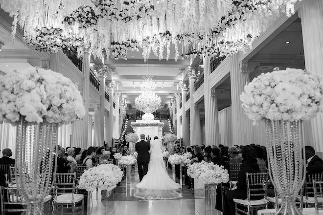 Father of the bride walking down the aisle with his daughter in a ballroom decorated in white florals. 