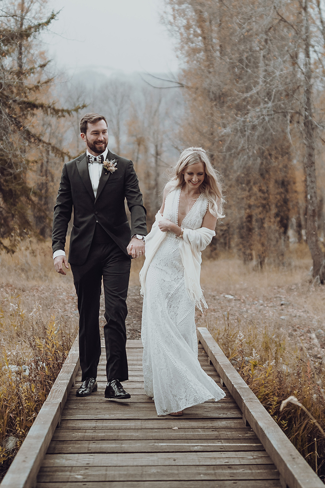 Groom holds hands with a smiling bride while walking across a wooden bridge at their autumn wedding in Jackson Hole, Wyoming. 