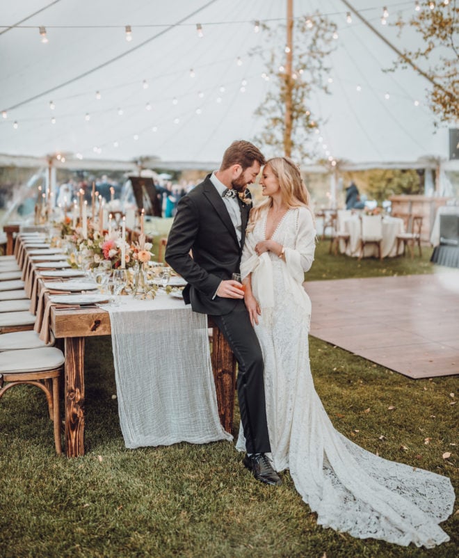 Groom scrunches his face and smiles at a smiling bride in long white gown and white shaw inside of a tent decorated with string lights, aspen trees and wooden guest tables set with lit candles and florals in a Jackson Hole, Wyoming.