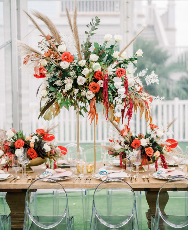 This gorgeous wedding décor features tall and short centerpieces filled with white and orange flowers, lush greenery, and pampas grass.