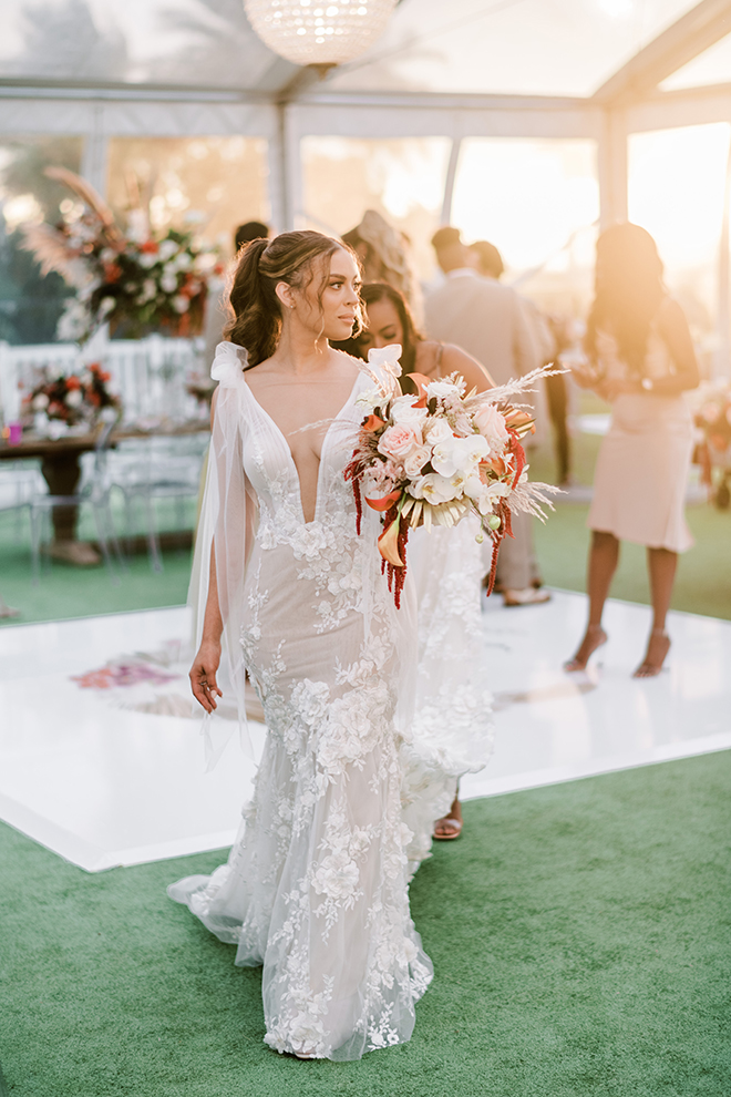 As the sun starts to set, the bride walks off the dance floor holding her bouquet as light illuminates her from behind. 