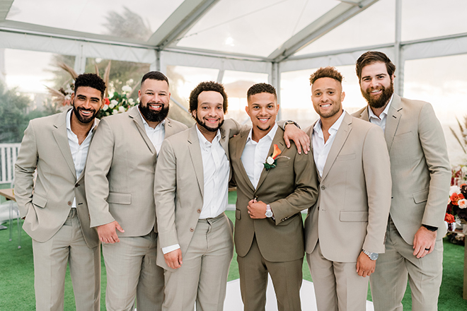 The groom in a taupe suit wears an orange boutonniere surrounded by groomsmen in light tan suits.